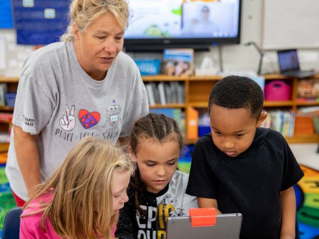  Teacher looks over three elementary students working on an iPad